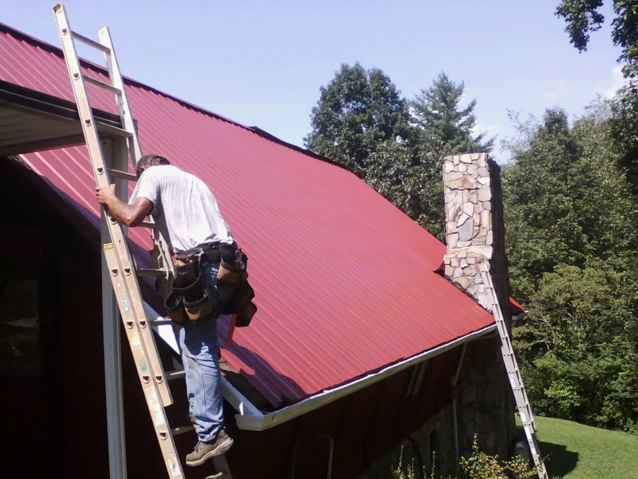 Roofer on a ladder beside a red metal roof and stone chimney in Seagrove
